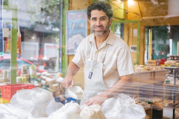 穀物を個人のバッグに詰めてカメラを見ている中年のラテン系穀物商が店のガラス越しに見える