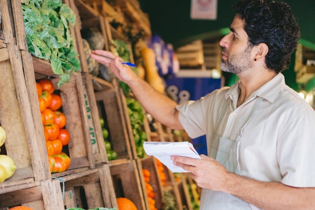 Middleaged Latin greengrocer writing down in a notebook what vegetables are needed and what is available