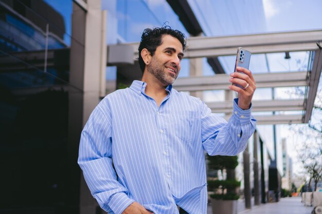 Middleaged Latin man in an oversized shirt making a video call on his smartphone in front of his office