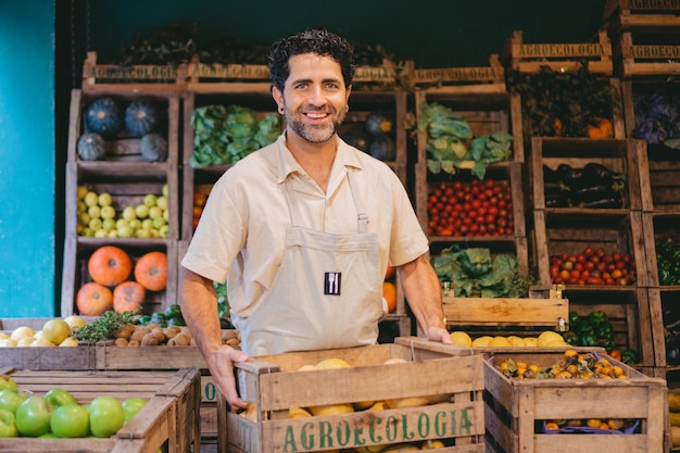 Middleaged Latin man smiling with a crate of pumpkins in a vegetable store while looking at the camera