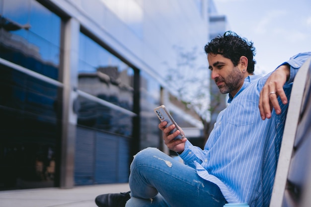 Латинский рабочий средних лет отдыхает на городской скамейке, проверяя свой мобильный телефон