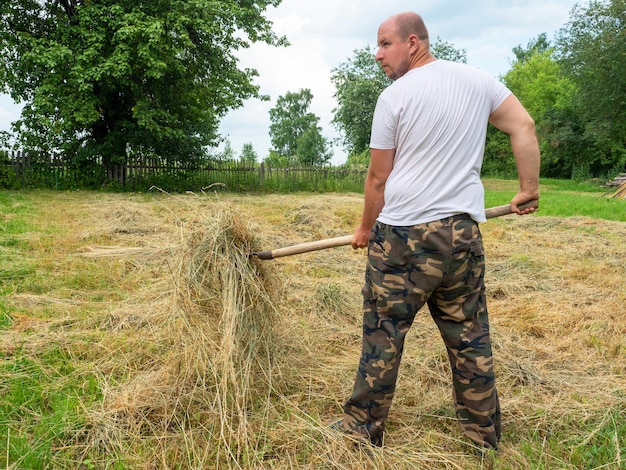 A middleaged man dressed in trousers and a Tshirt collects grass in a haystack with a fork The concept of working in rural areas harvesting livestock feed