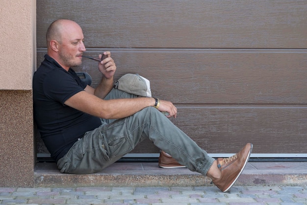 A middleaged man is resting on the sidewalk and smoking pipe\
portrait of caucasian bald man outdoors close up bad habits\
addiction unhealthy lifestyle concept copy space