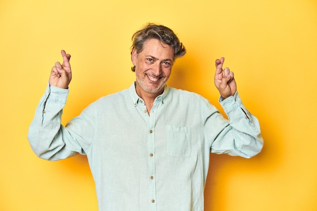 Middleaged man posing on a yellow backdrop crossing fingers for having luck