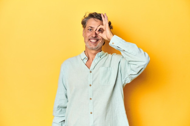 Middleaged man posing on a yellow backdrop excited keeping ok gesture on eye