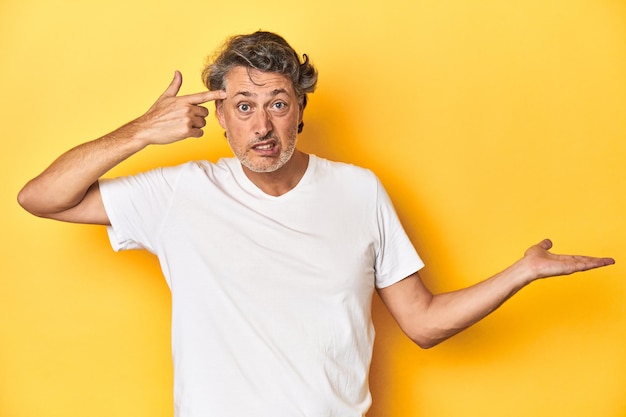 Middleaged man posing on a yellow backdrop holding and showing a product on hand