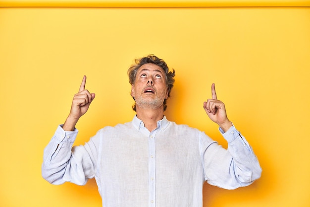 Middleaged man posing on a yellow backdrop pointing upside with opened mouth