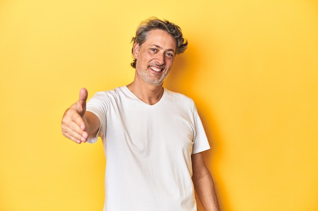 Middleaged man posing on a yellow backdrop stretching hand at camera in greeting gesture