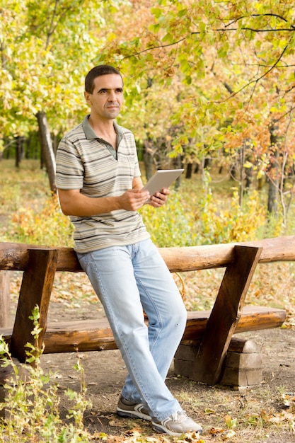 Middleaged man relaxing on a rustic wooden fence in woodland with a tablet in his hands