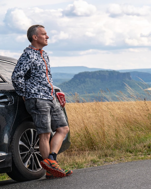 中年男性が車の近くに立って、ザクセン州スイスドイツの山の風景を眺める