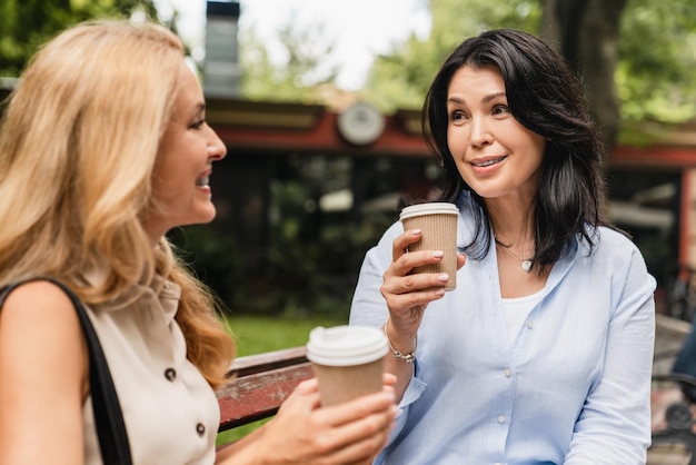 Middleaged mature caucasian sisters two mature women girlfriends best friends talking spending time together while sitting on the park bench drinking coffee in paper cups