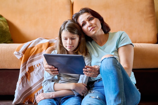 Middleaged mom and preteen daughter relaxing at home together with digital tablet
