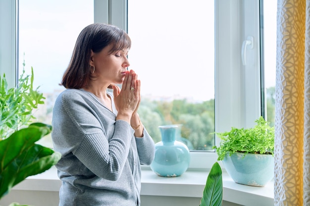 Middleaged serious woman praying near window at home Mature female folded her hands in prayer looking out window with eyes closed thinking experiencing suffering Faith hope religion soul