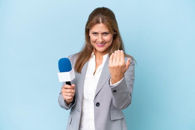 Middleaged TV presenter woman over isolated blue background inviting to come with hand Happy that you came