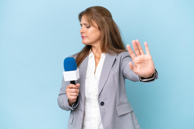 Middleaged TV presenter woman over isolated blue background making stop gesture and disappointed