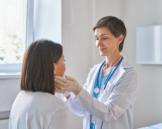 Middleaged woman doctor wearing gloves checking patients sore throat or thyroid glands