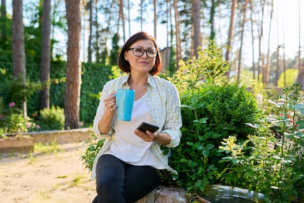 Женщина средних лет отдыхает в весеннем саду с чашкой чая и смартфоном в руках