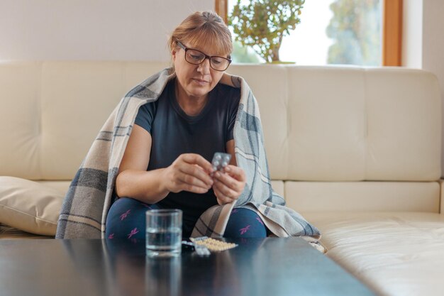 Middleaged woman holds a blister of pills and reads medical instructions sitting on the couch