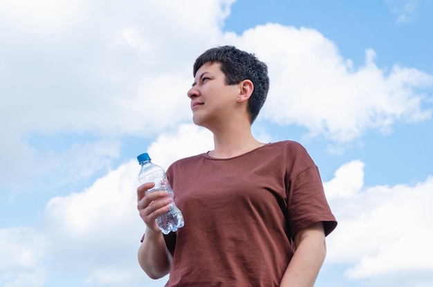 Женщина средних лет держит в руке пластиковую бутылку с водой на фоне ясного голубого неба с облаками