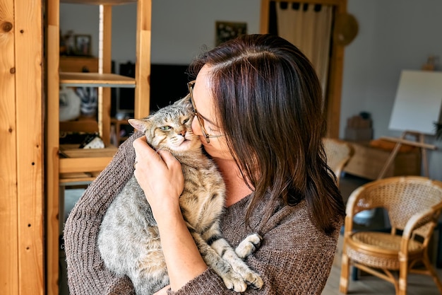 Middleaged woman kissing cute tabby cat in indoor scene Humananimal relationships Funny home pet Homeless pets