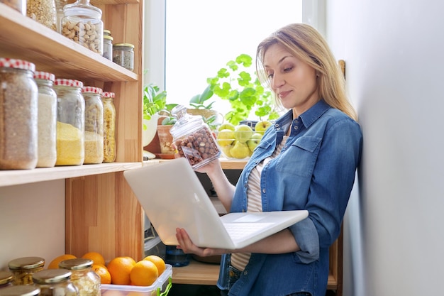 食品の缶を示すラップトップとパントリーのキッチンで中年の女性