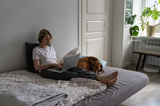 Женщина средних лет в пижаме ищет вакансию, печатая на ноутбуке, а рядом лежит собака