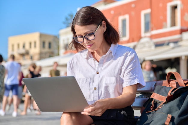 Женщина средних лет сидит на скамейке в городе с ноутбуком