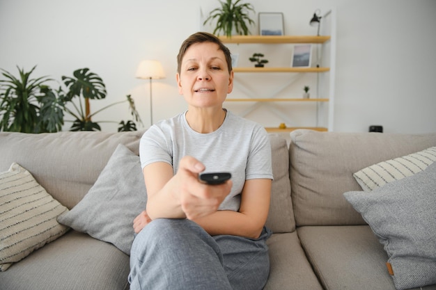 Женщина средних лет сидит на диване и включает телевизор пультом дистанционного управления