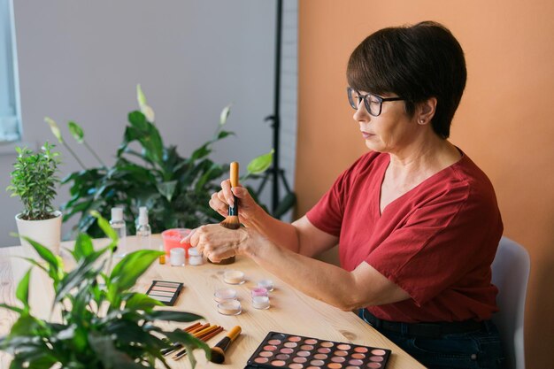 Middleaged woman talking on cosmetics with makeup eye shadows and blush palette and brushes while recording her video Mature female making video for her blog on cosmetics