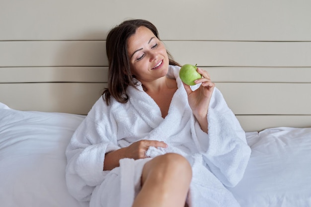 Middleaged woman in white bathrobe with green apple in bed