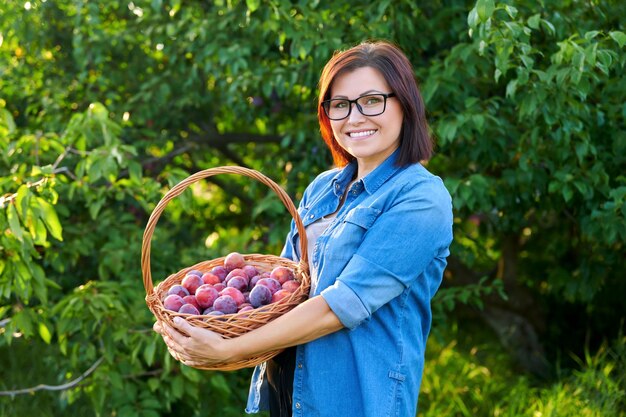 Middleaged woman with harvest of ripe plums in basket looking at camera