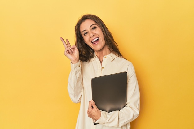 middleaged woman with laptop on yellow joyful and carefree showing a peace symbol with fingers