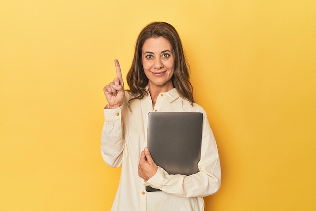 Middleaged woman with laptop on yellow showing number one with finger