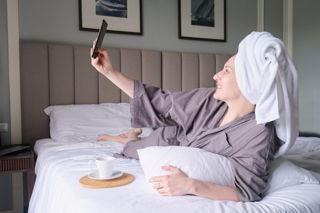 Middleaged woman with a towel on her head lies on the bed in the hotel room and takes a selfie