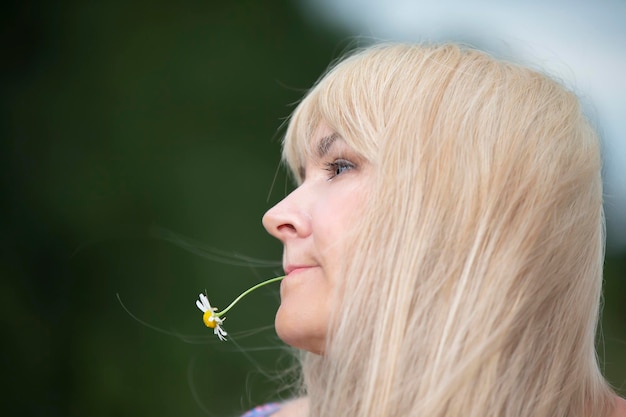 A middleaged woman with white hair holds a chamomile flower in her mouth