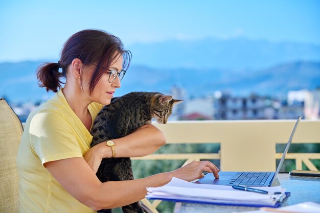 Middleaged woman working in home office on terrace with pet cat in her arms