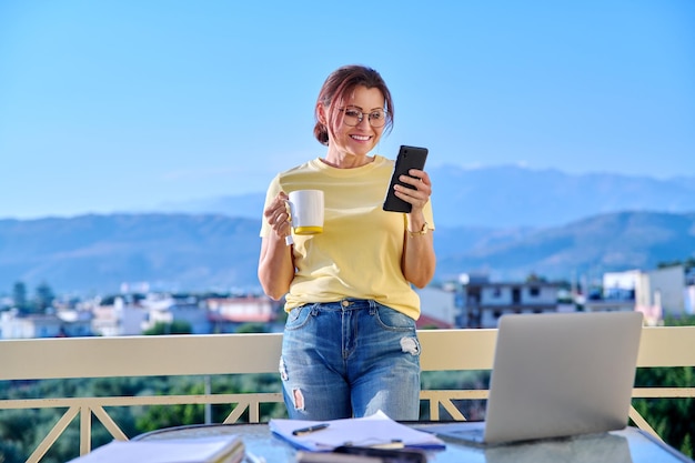 Middleaged woman working at home on terrace relaxing with mug of tea and smartphone