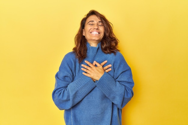 Middleaged woman on a yellow backdrop laughing keeping hands on heart concept of happiness