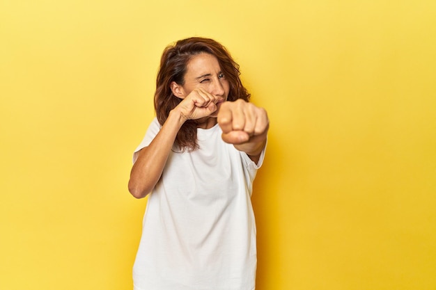 Middleaged woman on a yellow backdrop throwing a punch anger fighting due to an argument boxing