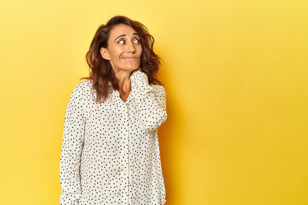 Photo middleaged woman on a yellow backdrop touching back of head thinking and making a choice