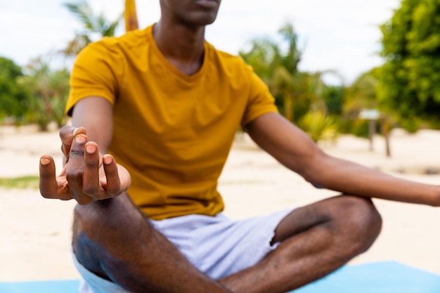 Midsection of biracial man practicing yoga meditation sitting on sunny beach. Lotus position, healthy lifestyle, wellbeing, relaxation, summer and vacation.