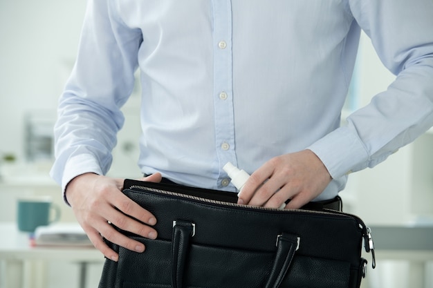 Midsection of businessman in formalwear putting sanitizer in small plastic bottle with spray into black leather handbag by his workplace