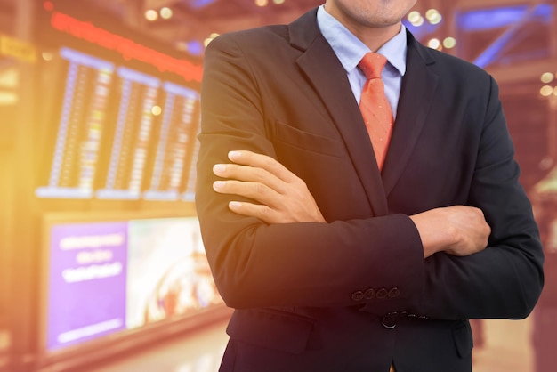 Midsection of businessman with arms crossed standing by arrival departure board at airport