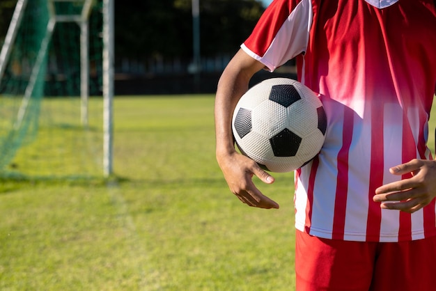 遊び場に立っているサッカーボールを持つ赤いジャージを着た白人の男性プレーヤーの中央部