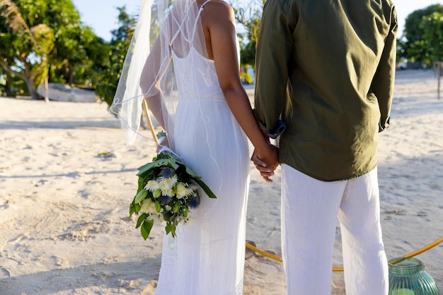 Midsection of caucasian newlywed couple holding hands and standing at beach at wedding ceremony. Unaltered, love, together, destination wedding, event, tradition, celebration, bouquet, hands, nature.