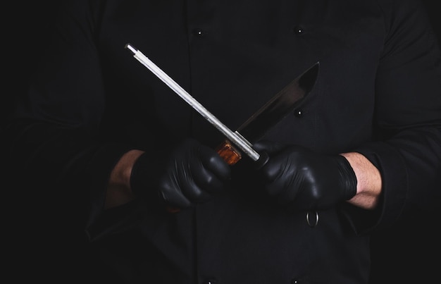 Foto sezione centrale di uno chef in uniforme che tiene un coltello da cucina a uno strumento sullo sfondo nero