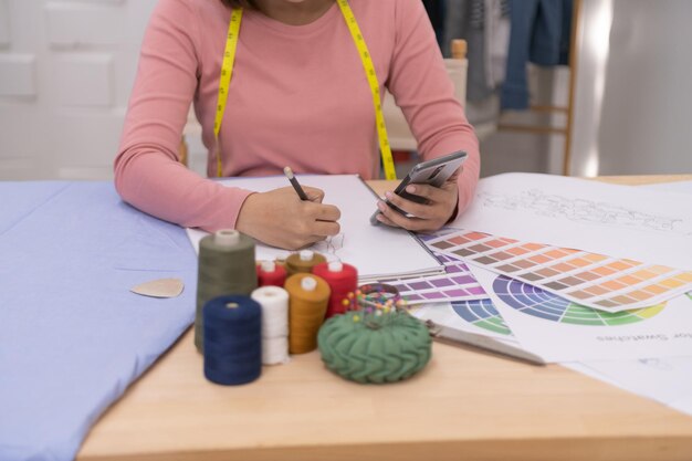 Midsection of female fashion designer drawing in book while using phone at desk