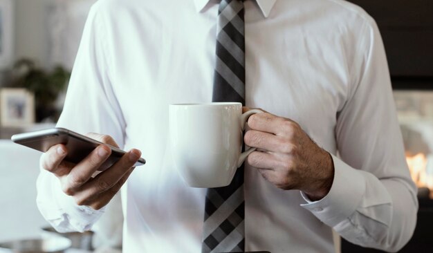 Фото Средняя часть бизнесмена, держащего чашку с кофе, использующего смартфон, стоя в офисе