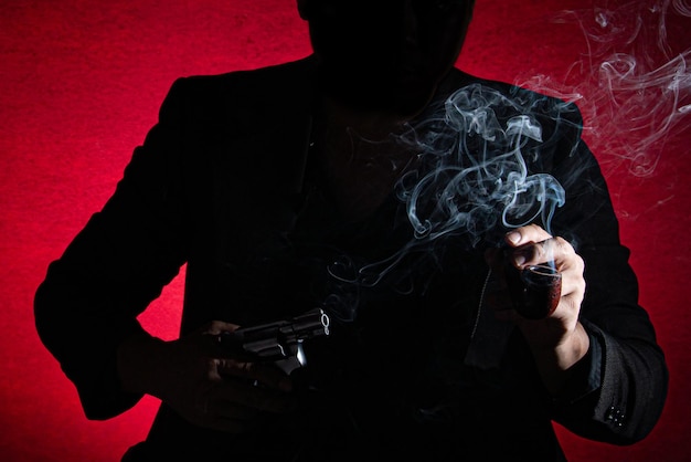 Фото Средняя часть силуэта мужчины, держащего курильную трубку и пистолет на красном фоне.