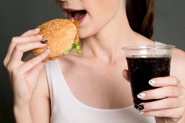 写真 灰色の背景の上にコーラを握りながらハンバーガーを食べている女性のミッドセクション